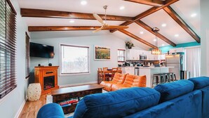 A living room with a blue sofa, wooden beams, a fireplace, and a kitchen in the background. - Greek Spa • Private Acreage • Stonefire pizza oven (Front Royal)