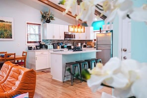 Una cocina moderna con armarios blancos, una isla central con taburetes, un comedor con mesa y sillas de madera, y un amplio ventanal con persianas.