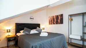 A neatly made bed with a dark quilt, two bedside lamps, a wall-mounted picture, and a shelf with a folded towel. - Adriatic View Apartment by DuHomes (Dubrovnik)