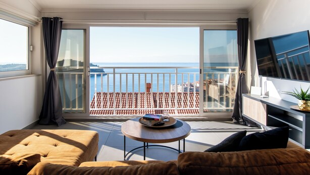 A modern living room with a balcony overlooking the sea, a flat-screen TV, and a round coffee table with books. - Adriatic View Apartment by DuHomes (Dubrovnik)