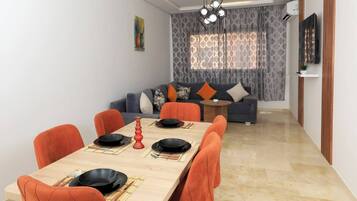 Un espace repas moderne avec une table rectangulaire, des chaises recouvertes d’un tissu orange et un canapé en arrière-plan.