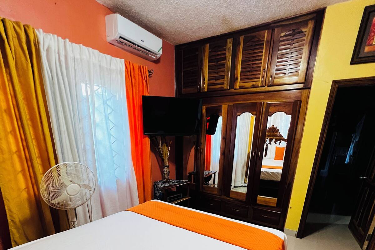 Una habitación de hotel con una cama, cortinas, un ventilador, un televisor y una puerta que da a otra habitación.