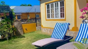 Een zonnig terras met twee loungestoelen en een kleine tafel, omgeven door een verzorgd gazon en een geel gebouw met een raam.