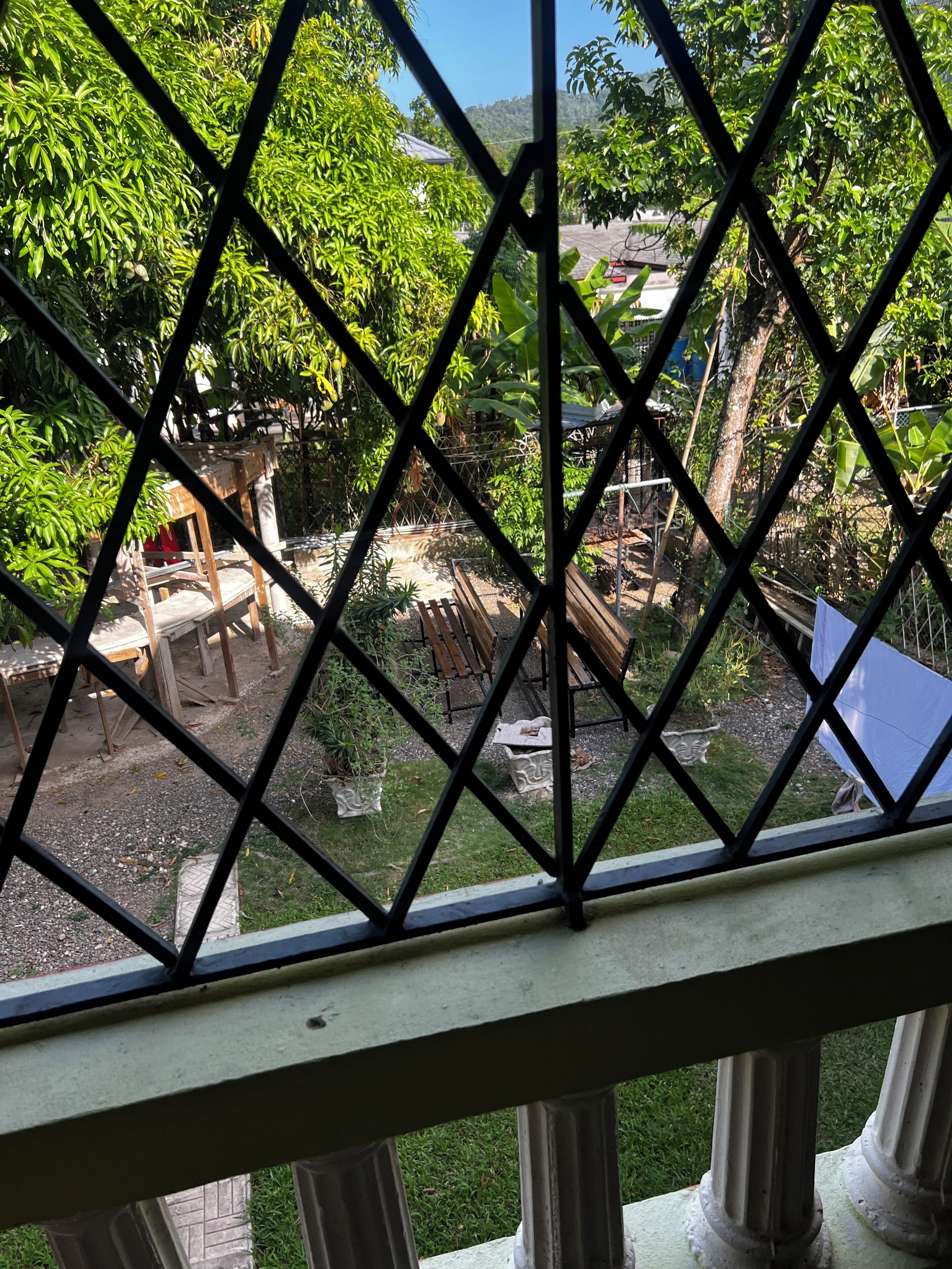 Vista a través de una ventana con diseño de rombos de un jardín con un banco de madera, una mesita y diversas plantas en macetas.