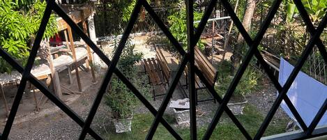 Vista a través de una ventana con diseño de rombos de un jardín con un banco de madera, una mesita y diversas plantas en macetas.