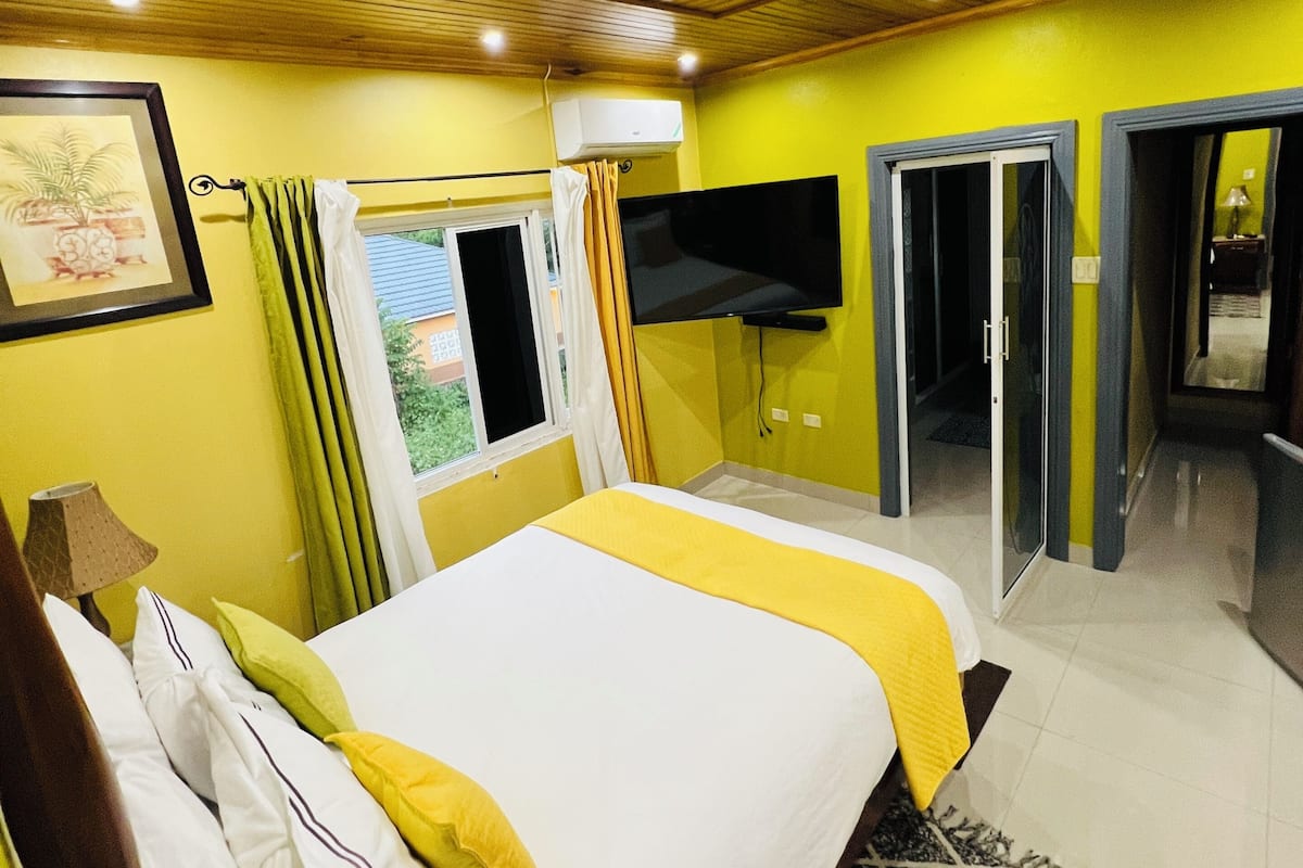 Habitación de hotel con techo de madera, cama con ropa de cama blanca y amarilla, televisor de pantalla plana y ventana con cortinas.