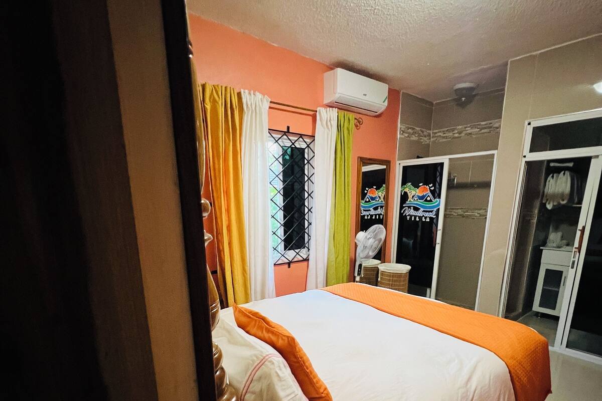 Habitación de hotel con cama, cortinas, unidad de aire acondicionado, ventana con reja de metal y puerta corredera que da al baño.