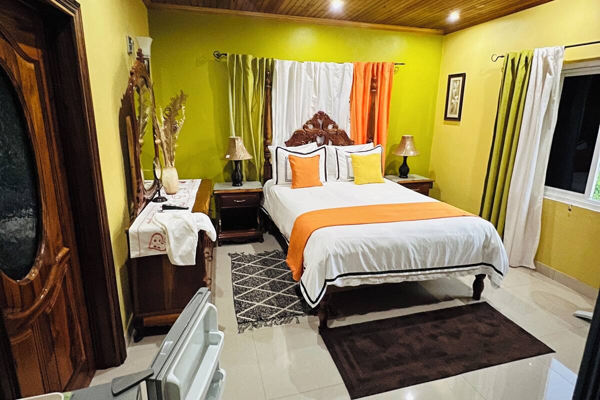 Un dormitorio con techo de madera, una cama con ropa de cama blanca y detalles naranja, una mesita de noche con lámpara, una ventana con cortinas y una puerta.