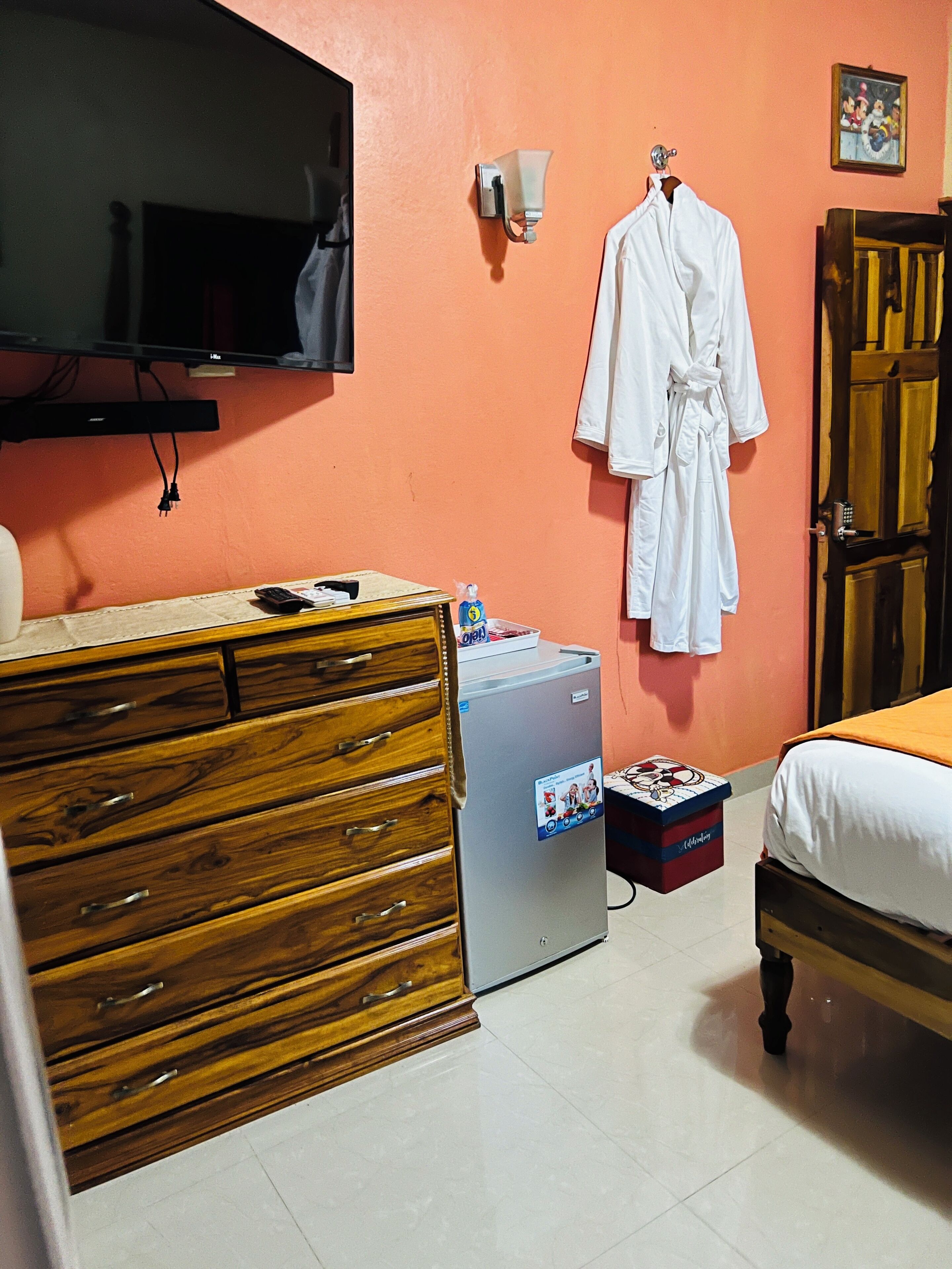 A hotel room with a wooden dresser, a small refrigerator, a bed with an orange blanket, a white robe hanging on the wall, and a television mounted above the dresser.