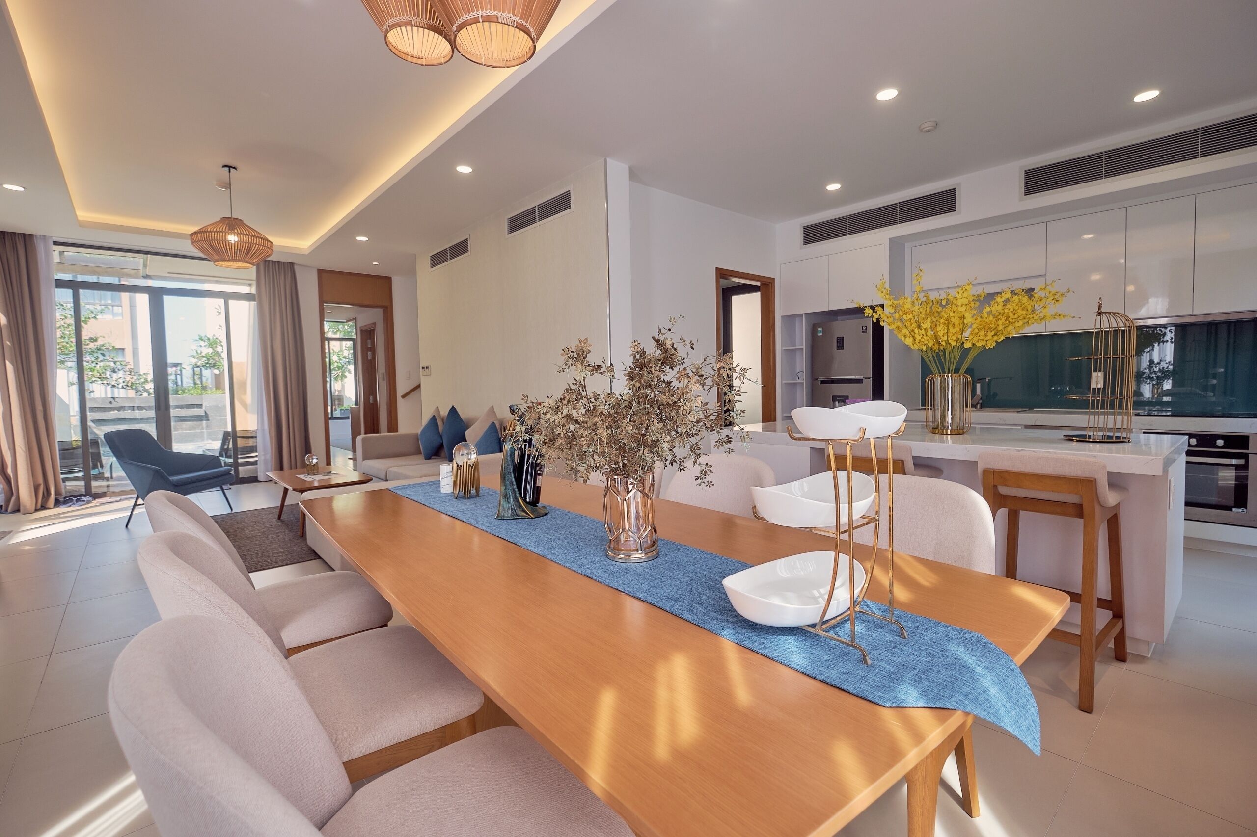 Uma sala de estar moderna com uma longa mesa de madeira, assentos confortáveis, área de cozinha e vista através de amplas janelas.