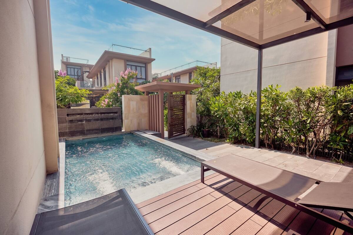 Ein Schwimmbecken mit Holzterrasse und Liegestühlen, umgeben von Grünflächen und modernen Gebäuden.