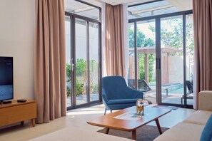 A modern living room with a television, a blue armchair, a wooden coffee table, and large glass doors leading to a patio with outdoor furniture. - Sunrise Pool Villa Cam Ranh  (Cam Lam)