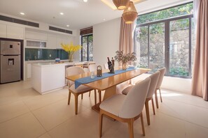A modern kitchen with a central island, dining table with chairs, and large windows with curtains. - Sunrise Pool Villa Cam Ranh  (Cam Lam)