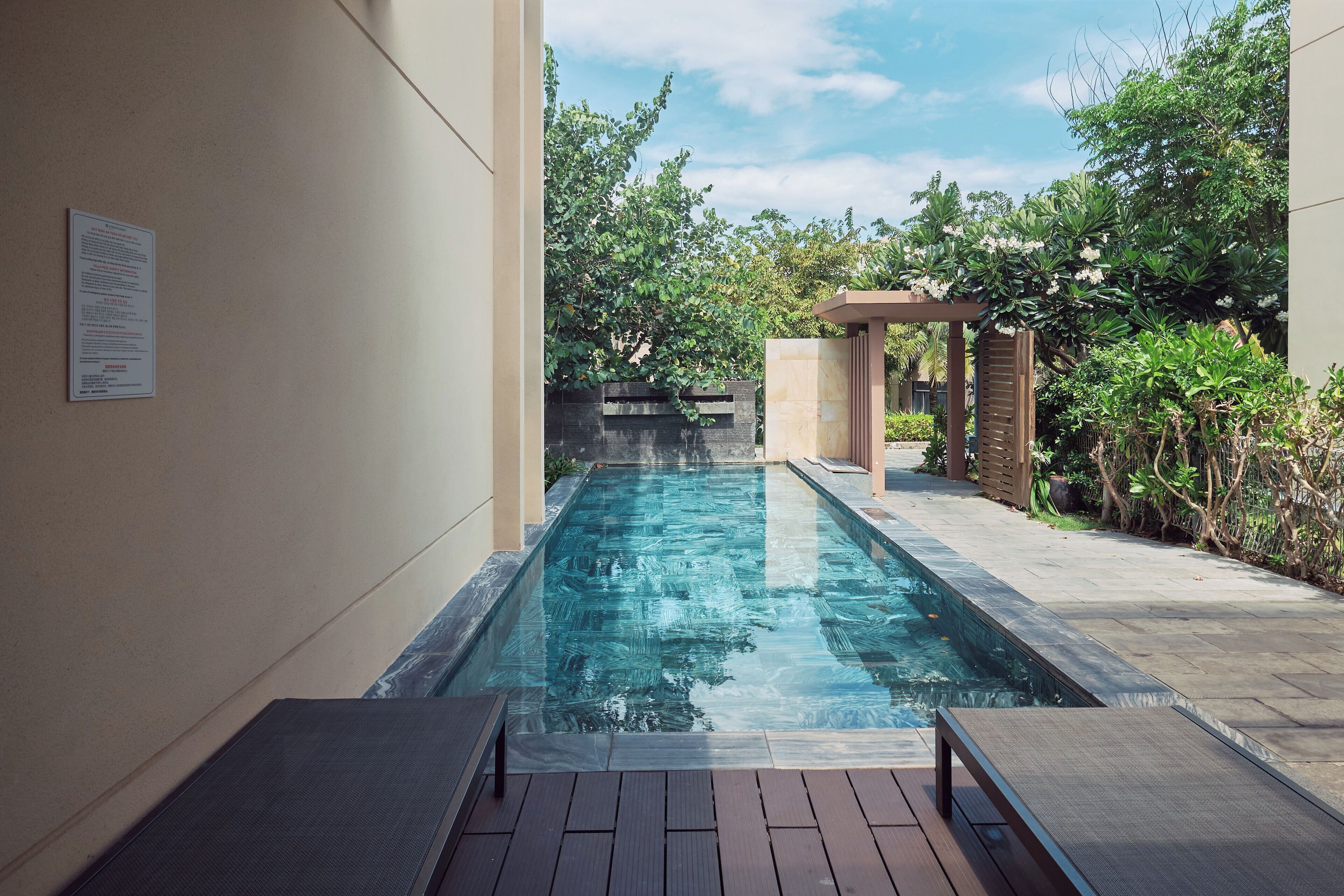 A swimming pool with a wooden deck, surrounded by greenery and a pergola.