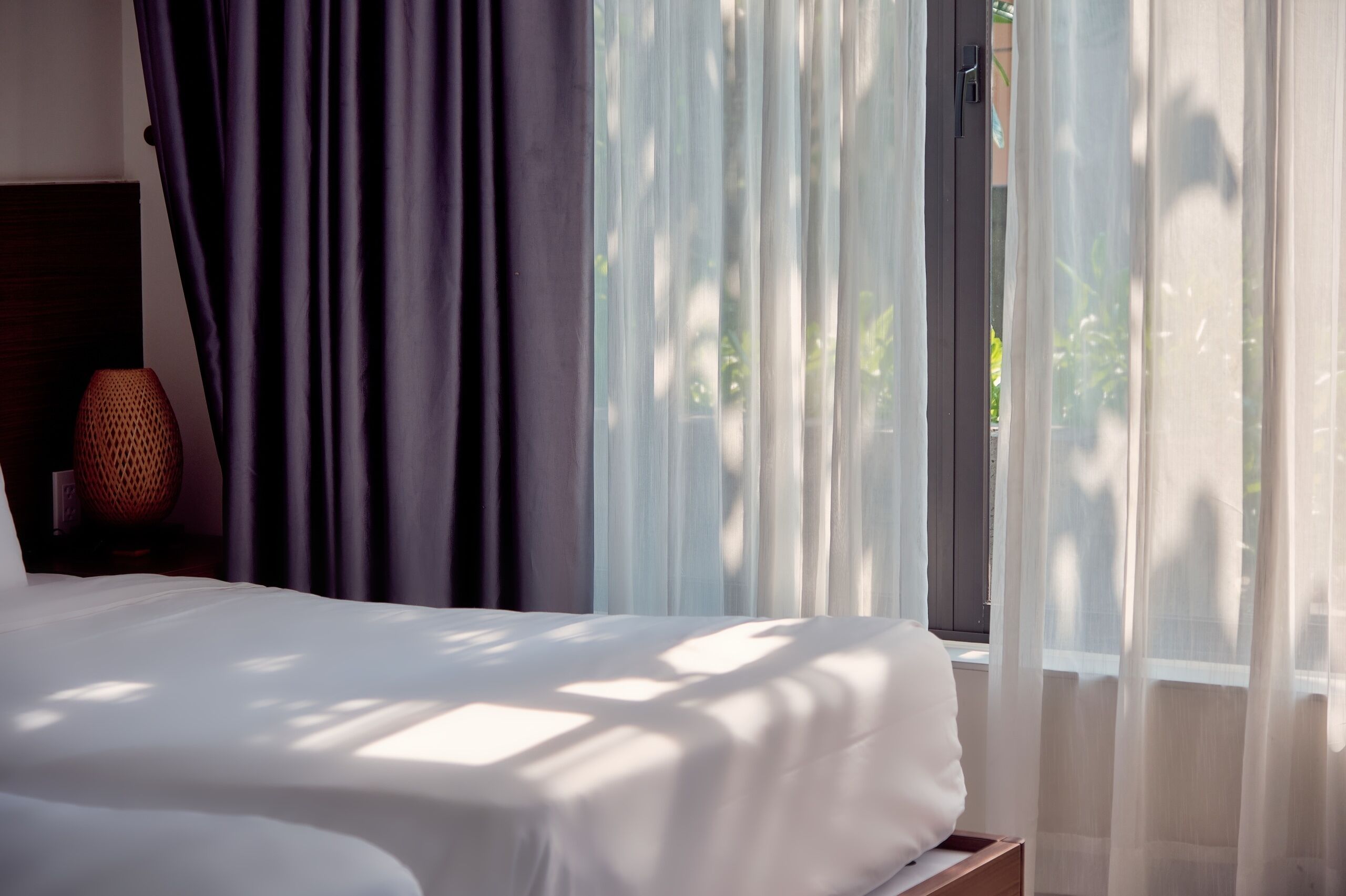 A bedroom with a bed, a nightstand, and a window with sheer curtains.