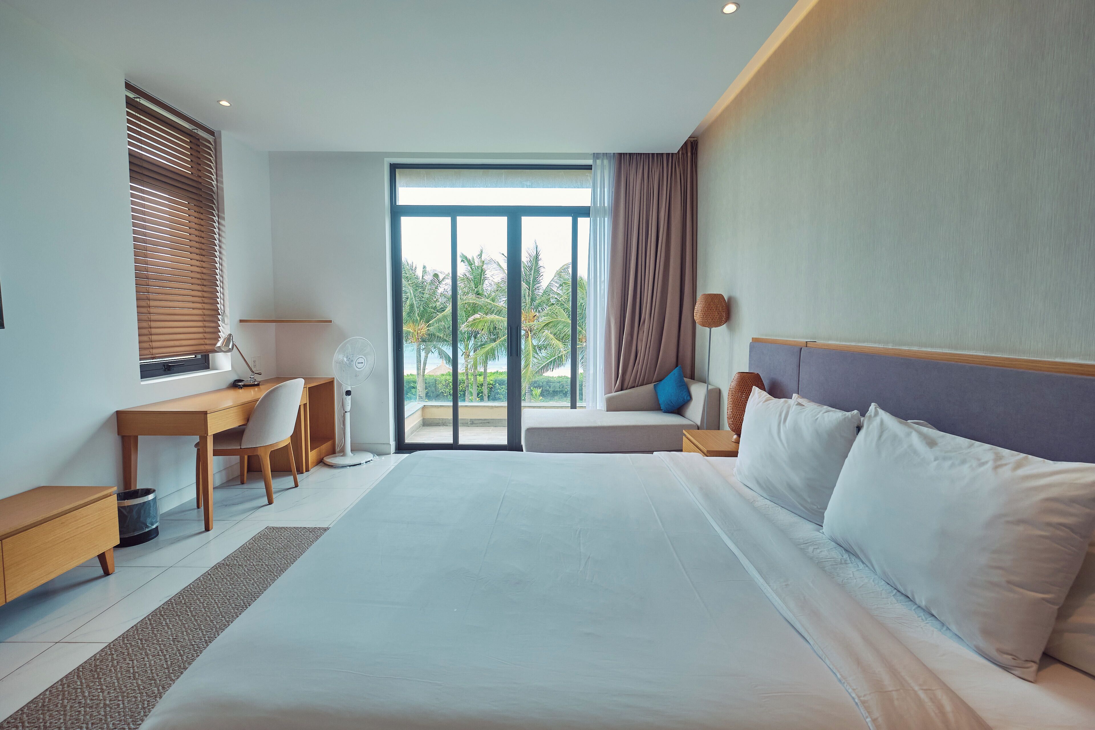 A hotel room with a large bed, a desk, a chair, a laptop, a lamp, and a view of palm trees through the window.