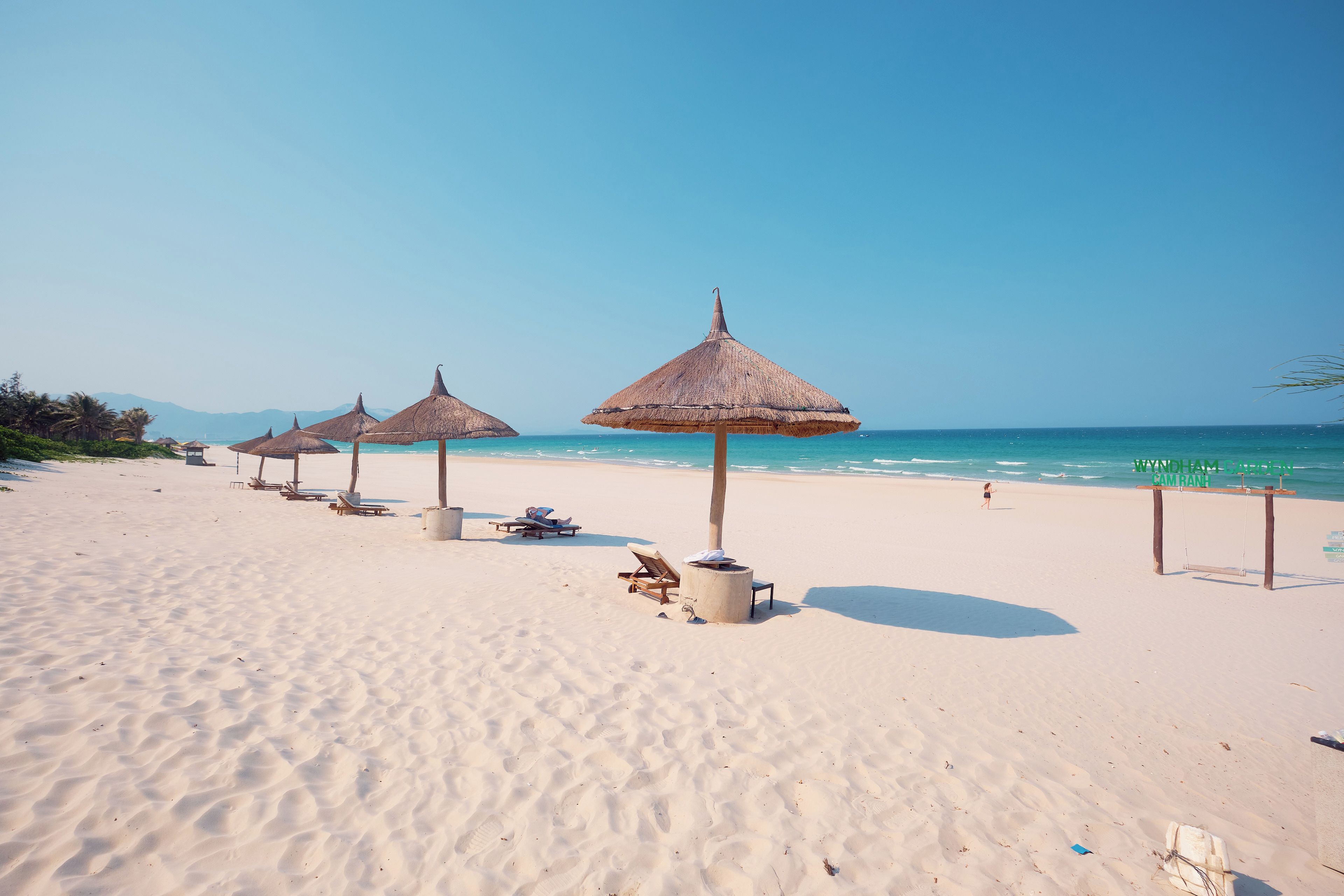 A beach with thatched umbrellas and lounge chairs, a clear sky, and calm sea.