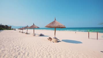 A beach with thatched umbrellas and lounge chairs, a clear sky, and calm sea.