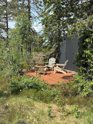 Eine Holzterrasse mit Adirondack-Stühlen und einer Bank, umgeben von dichtem Grün und Bäumen.