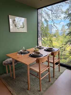 A dining area with a table set for two, a framed picture on the wall, and a view of trees and sky through a large window.