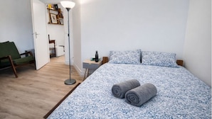 Un dormitorio con cama, mesita de noche, lámpara de pie, una silla y una puerta que da a otra habitación.