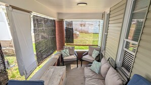 A covered patio with a wooden table, two blue chairs, a beige sofa with cushions, and a view of a backyard.