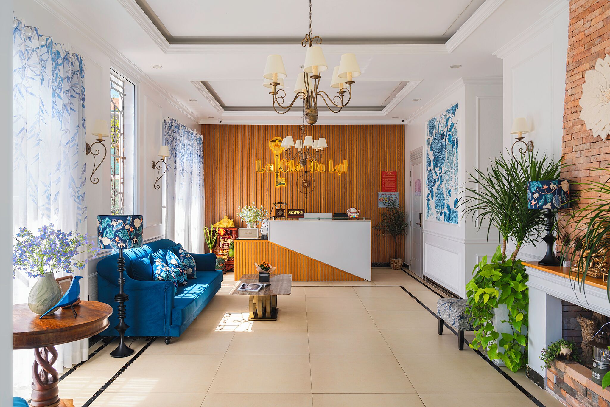A hotel lobby with a reception desk, a blue sofa, a chandelier, and decorative plants.