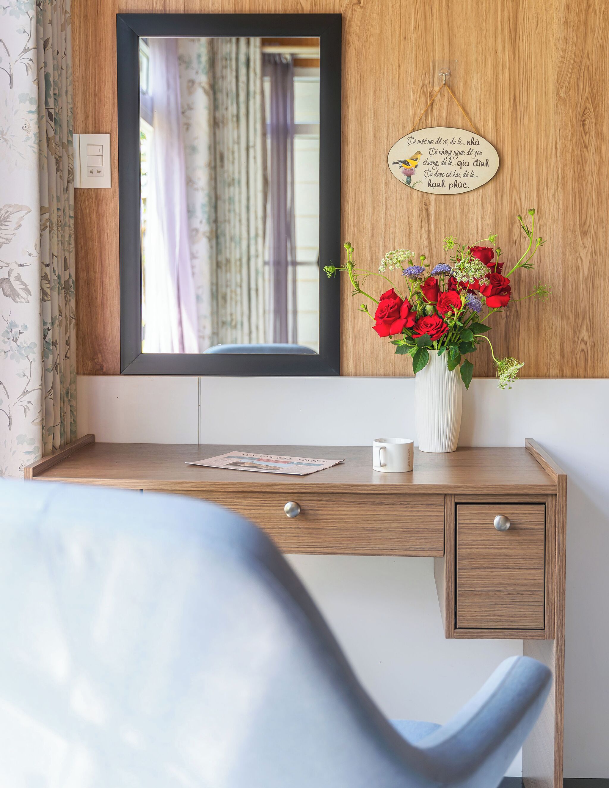 Un bureau en bois avec un miroir, un vase de roses rouges, un journal et une tasse blanche.