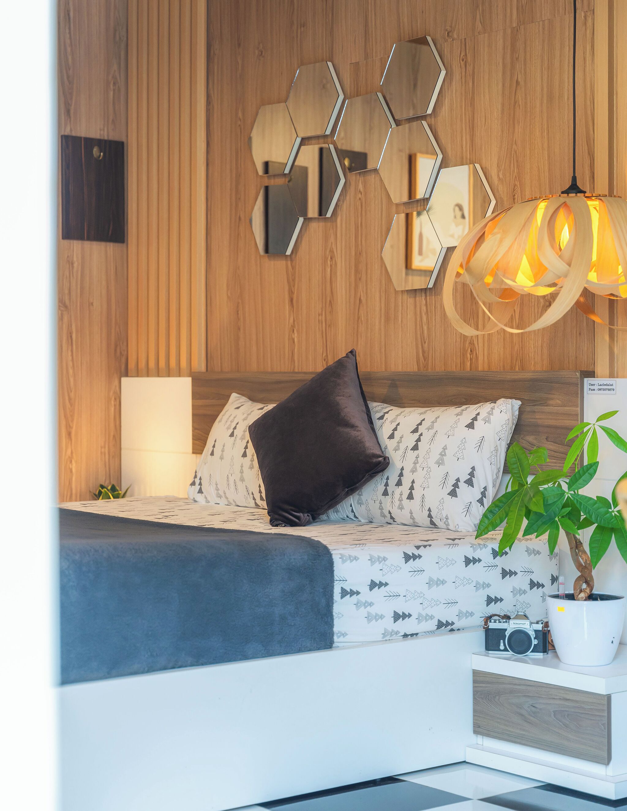 Un dormitorio moderno con cabecera de madera, espejo de pared con diseño geométrico, lámpara de noche, almohadas estampadas, una planta en maceta y una pequeña mesa con una cámara.