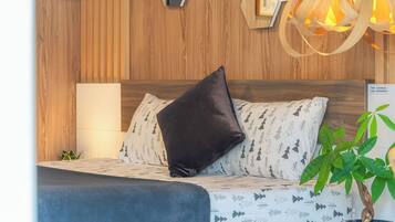 Un dormitorio moderno con cabecera de madera, espejo de pared con diseño geométrico, lámpara de noche, almohadas estampadas, una planta en maceta y una pequeña mesa con una cámara.