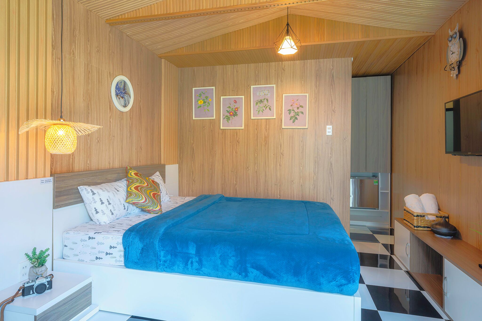 A bedroom with a bed, a bedside table with a camera, a lamp, and framed pictures on the wall.