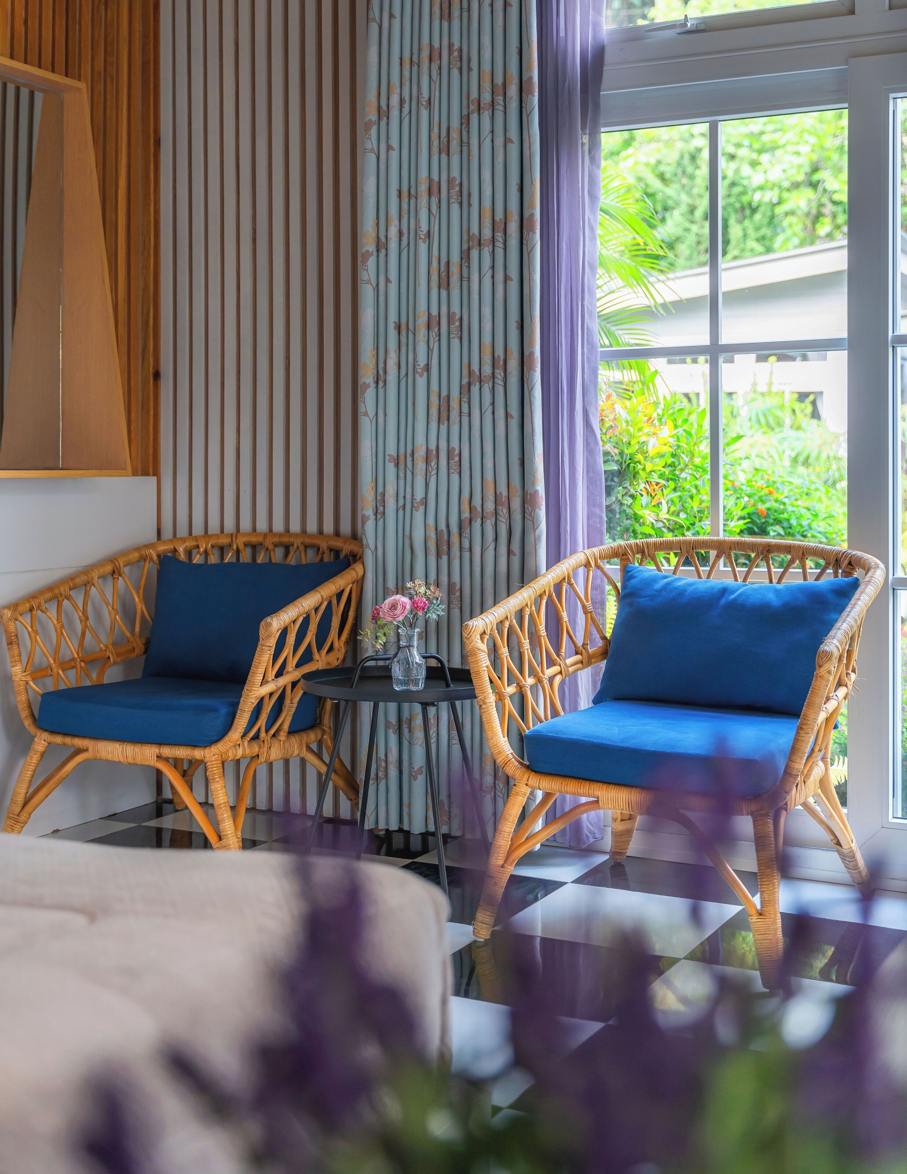 A room with two wicker chairs with blue cushions, a small table with a vase of flowers, and a window with a view of greenery.