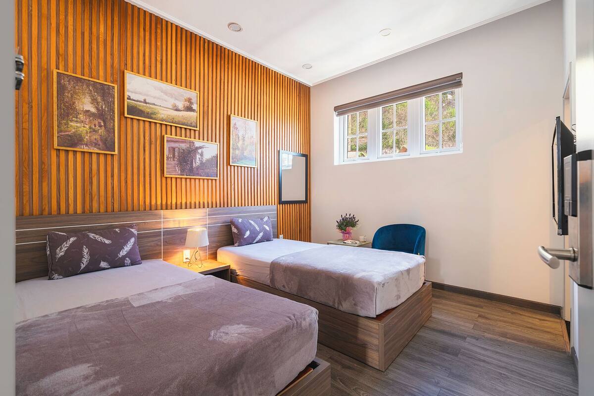 A modern hotel room with two beds, wooden paneling on the walls, and a large window providing natural light.