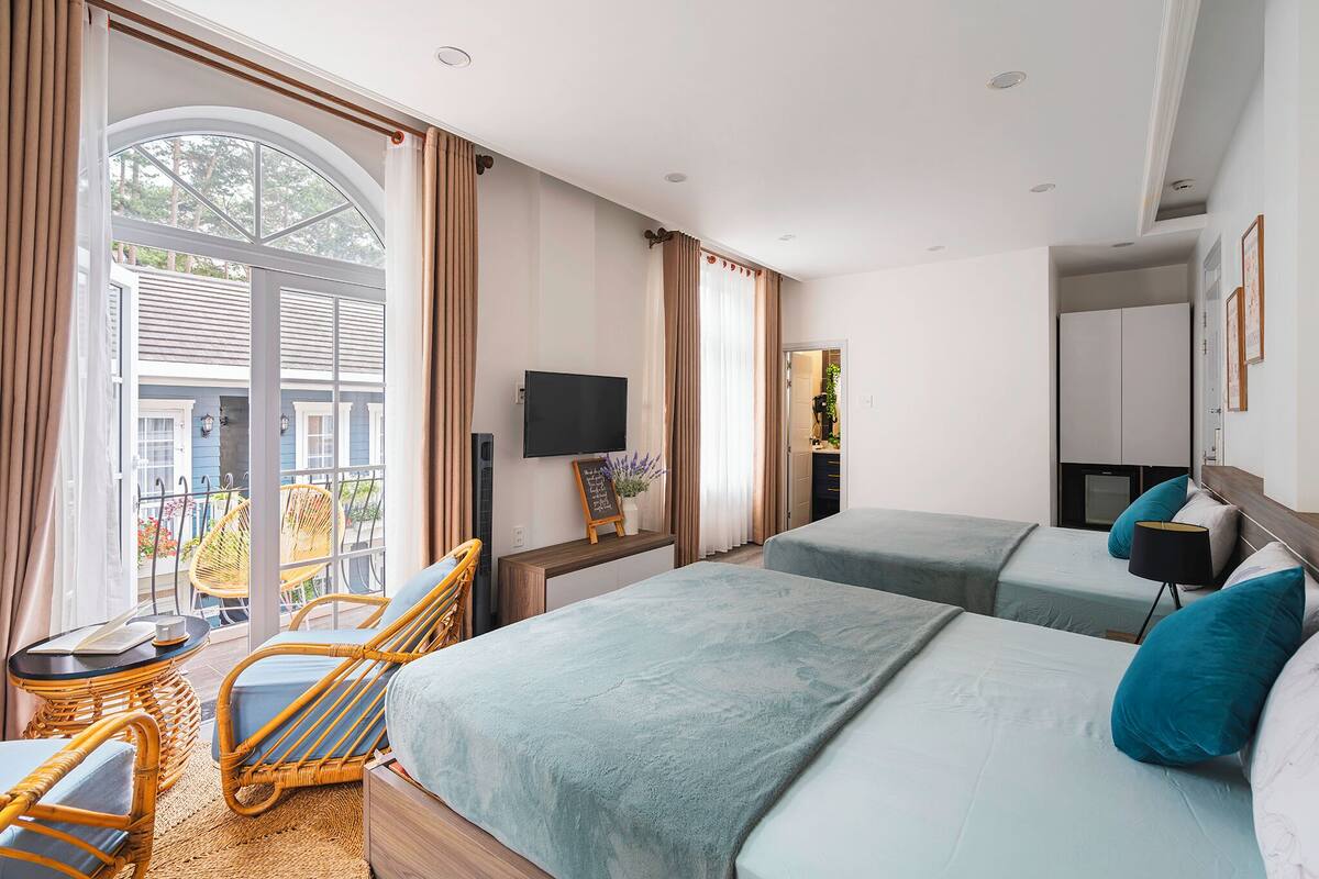 A hotel room with two beds, a TV, a balcony with wicker chairs, and a view of the outside.