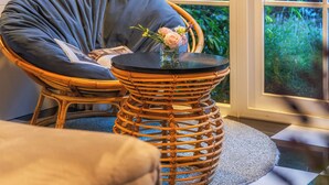 Une pièce avec un fauteuil en osier, une petite table ronde ornée d’un vase de fleurs, une vue sur la verdure à l’extérieur et un tapis à motifs sur le sol.