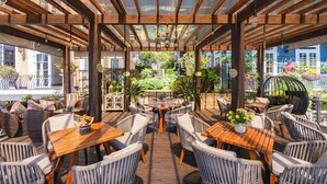 Une terrasse extérieure avec des tables en bois et des chaises en osier, entourée d’une pergola en bois agrémentée de guirlandes lumineuses.