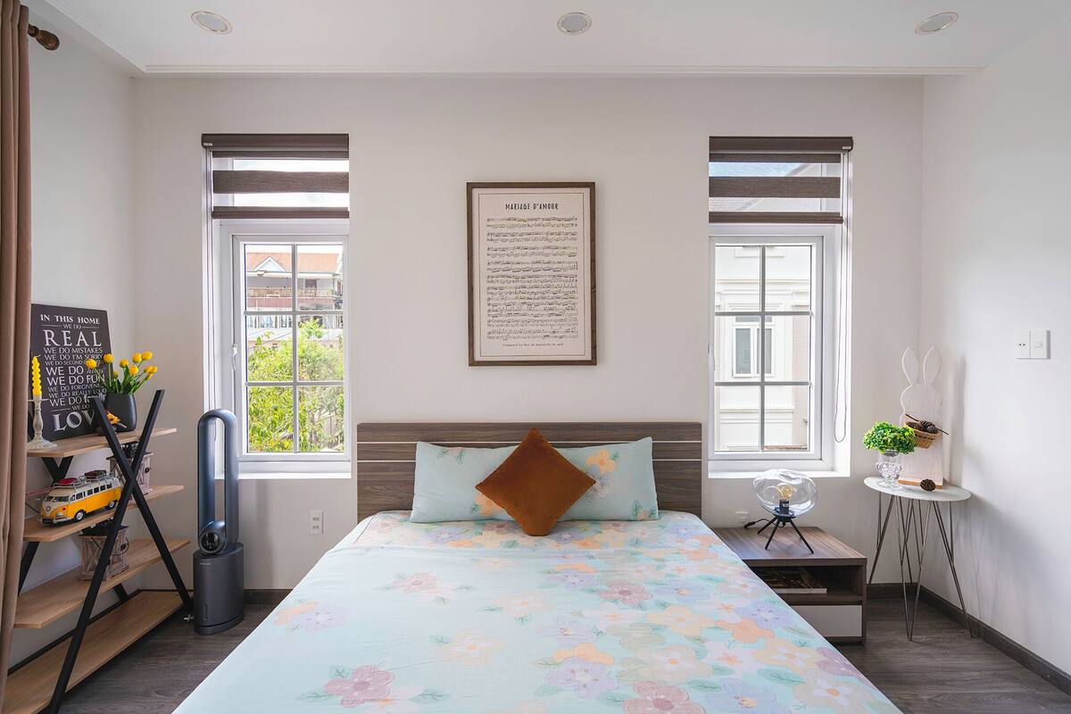 A bedroom with a bed, a bedside table, a standing fan, a window with blinds, a framed quote on the wall, and a small plant on a side table.