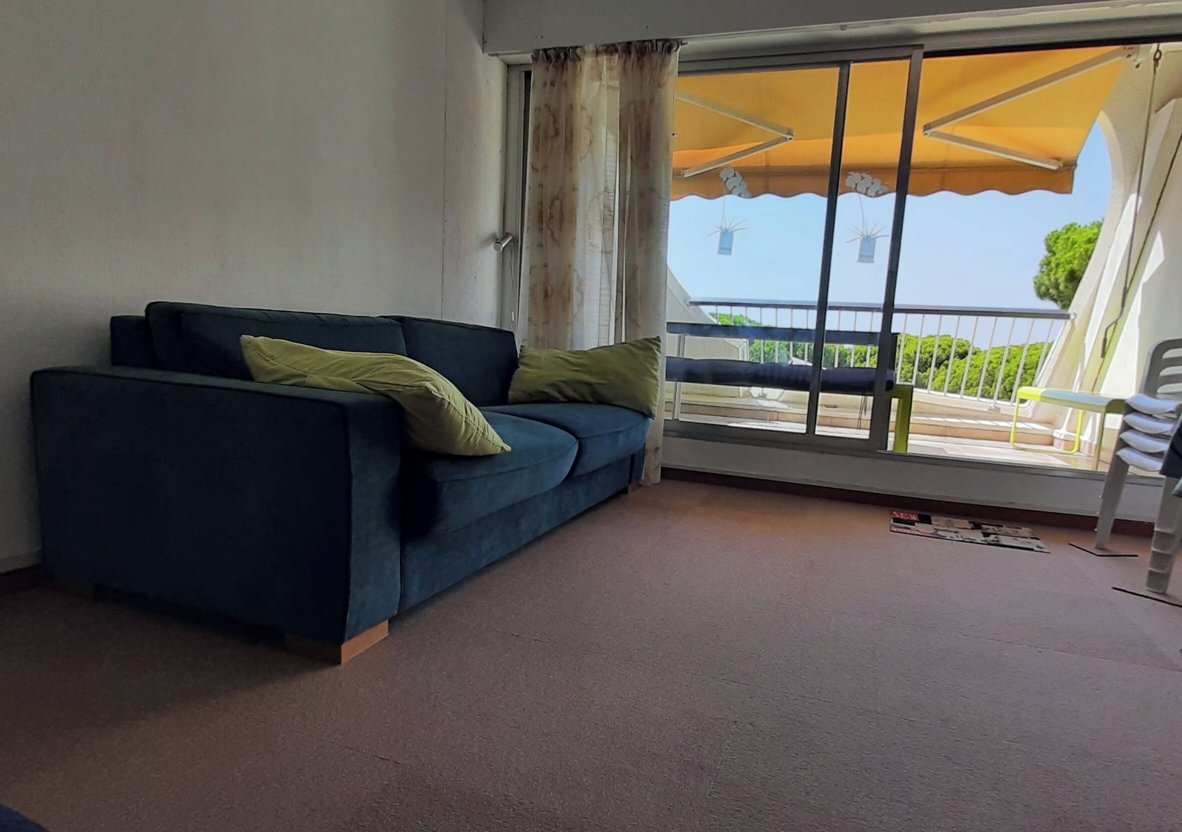 Une pièce avec un canapé bleu foncé, deux coussins verts, une grande fenêtre surmontée d’un auvent jaune et un balcon doté d’une balustrade et d’une plante suspendue.