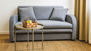 Ein graues Sofa mit zwei Kissen, ein gewebter Teppich, ein kleiner runder Tisch mit einem gewebten Korb und einem gelben Behälter, und ein goldener Vorhang.