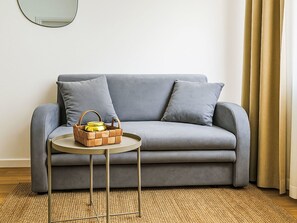 En grå soffa med två kuddar, en vävd matta, ett litet runt bord med en vävd korg och en gul behållare samt en guldfärgad gardin.