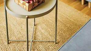 Ein Zimmer mit einem Bett, einem kleinen runden Tisch mit einem Korb, der ein Glas und Blumen enthält, und einem Teppich auf dem Boden.