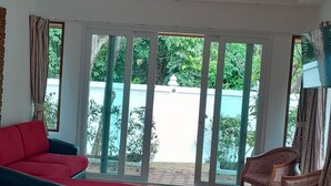 A room with a wooden floor, a bed, a red sofa, a wooden chair, and a large window with a view of greenery.