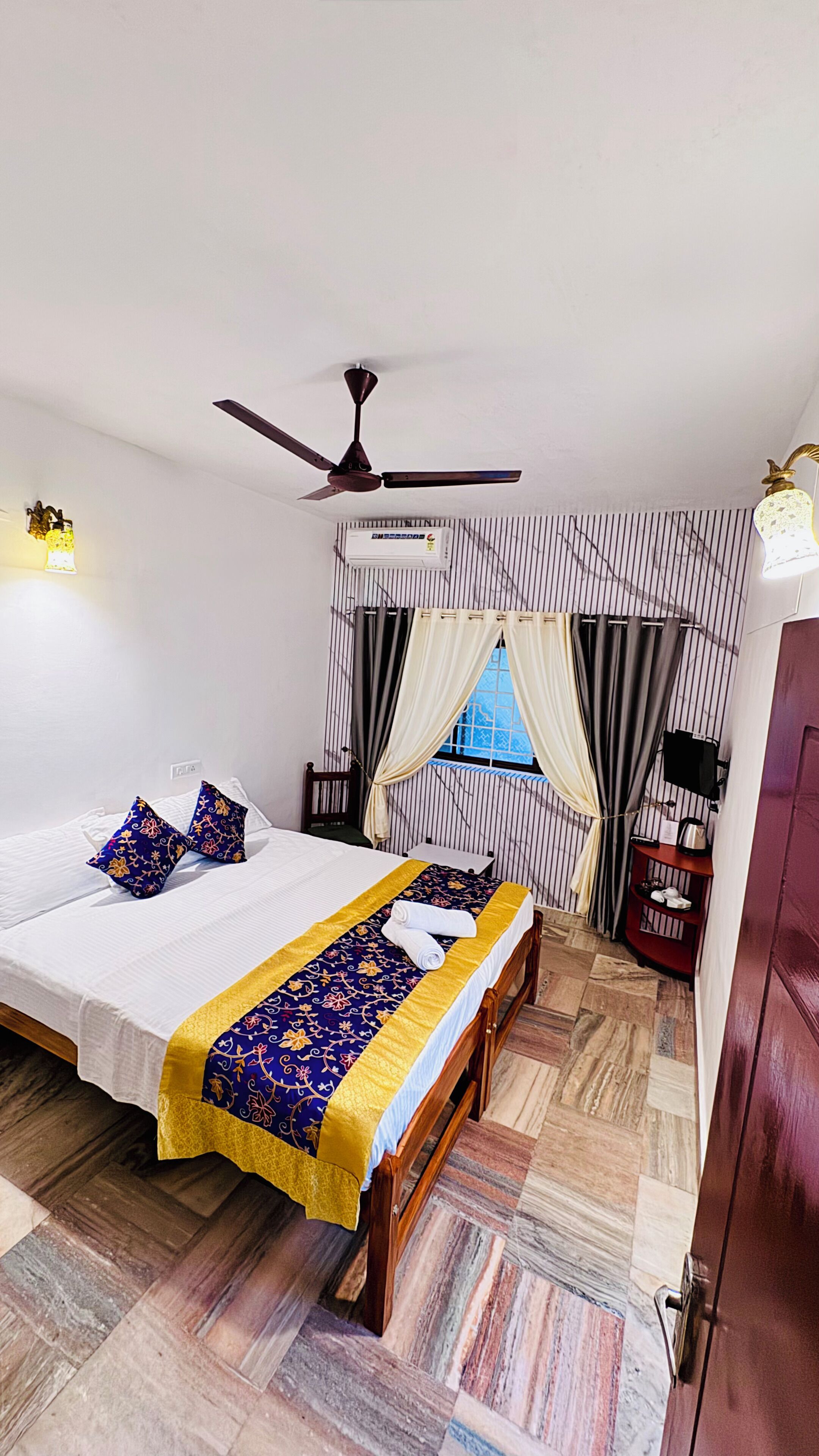 Une chambre à coucher comprenant un lit, un ventilateur de plafond, une fenêtre avec des rideaux, une table de chevet et une lampe fixée au mur.