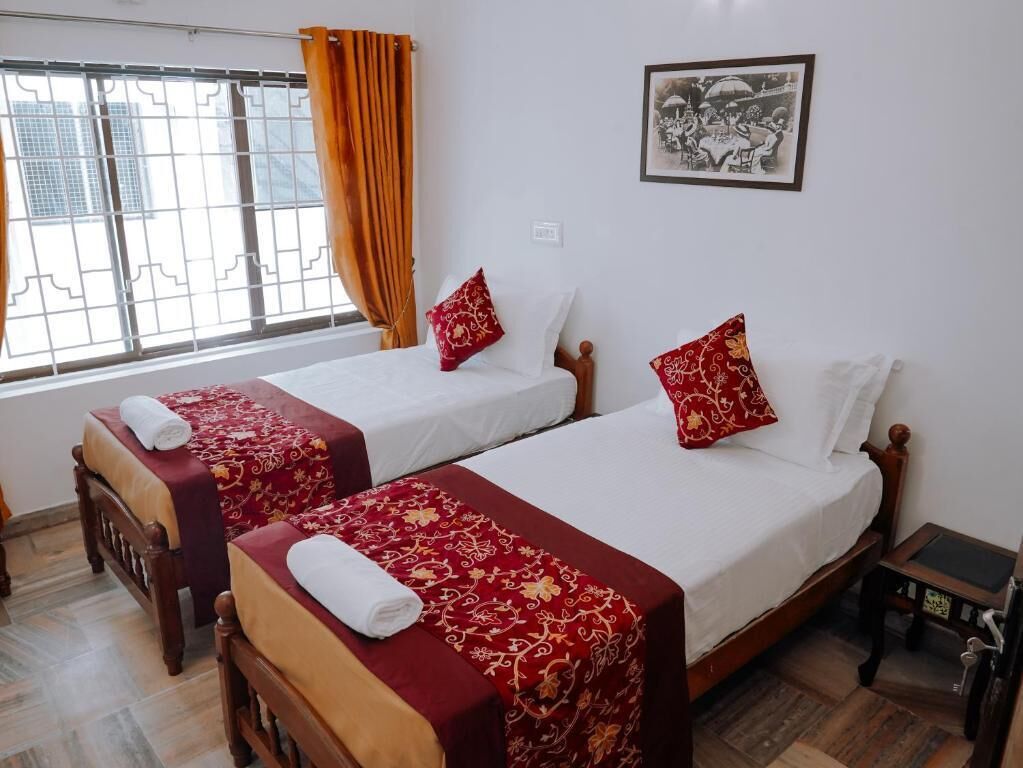 Habitación de hotel con dos camas individuales, cada una con una colcha y almohadas estampadas en rojo y dorado. Se ve una ventana con cortinas naranjas, un cuadro en la pared y una pequeña mesa de madera con superficie negra.