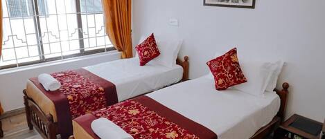 Habitación de hotel con dos camas individuales, cada una con una colcha y almohadas estampadas en rojo y dorado. Se ve una ventana con cortinas naranjas, un cuadro en la pared y una pequeña mesa de madera con superficie negra.