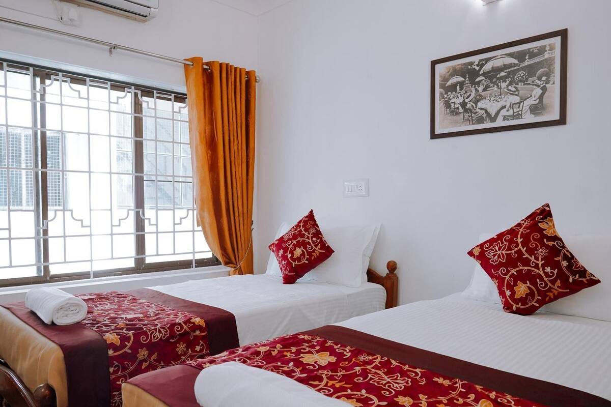 A hotel room with two beds, each with red and white bedding, a window with orange curtains, and a framed picture on the wall.