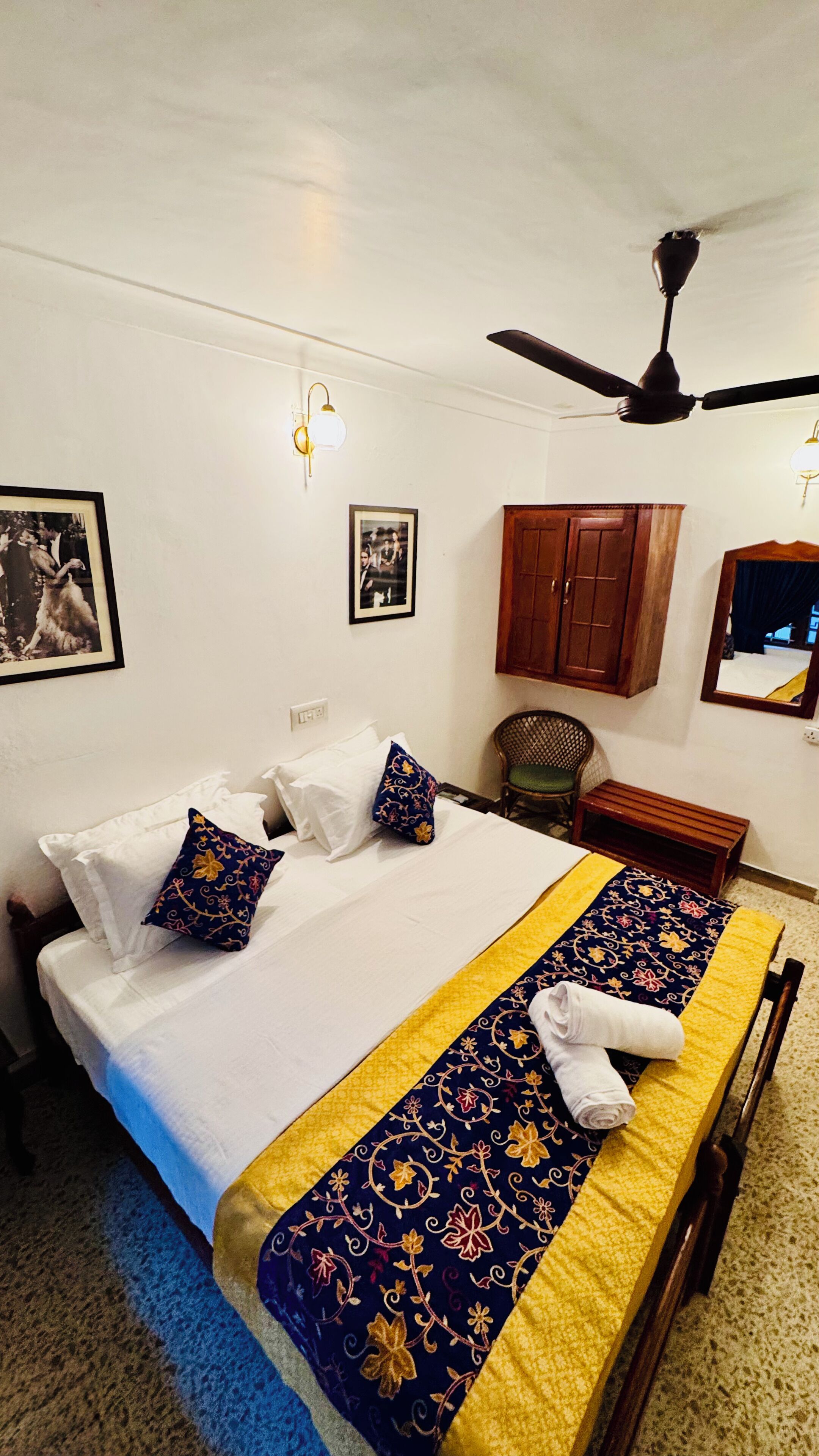 Cama bem arrumada com colcha azul e amarela, cabeceira de madeira e um ventilador de teto.