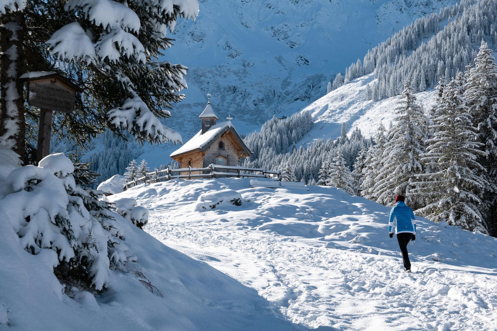 A snow-covered mountain path leading to a wooden cabin with a bell tower, surrounded by snow-laden trees and a clear sky.
