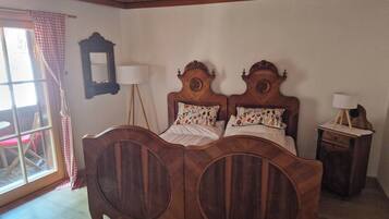 Une chambre avec un lit en bois doté de deux têtes de lit rondes, une table de chevet avec une lampe, un miroir au mur et une fenêtre avec des rideaux à carreaux.