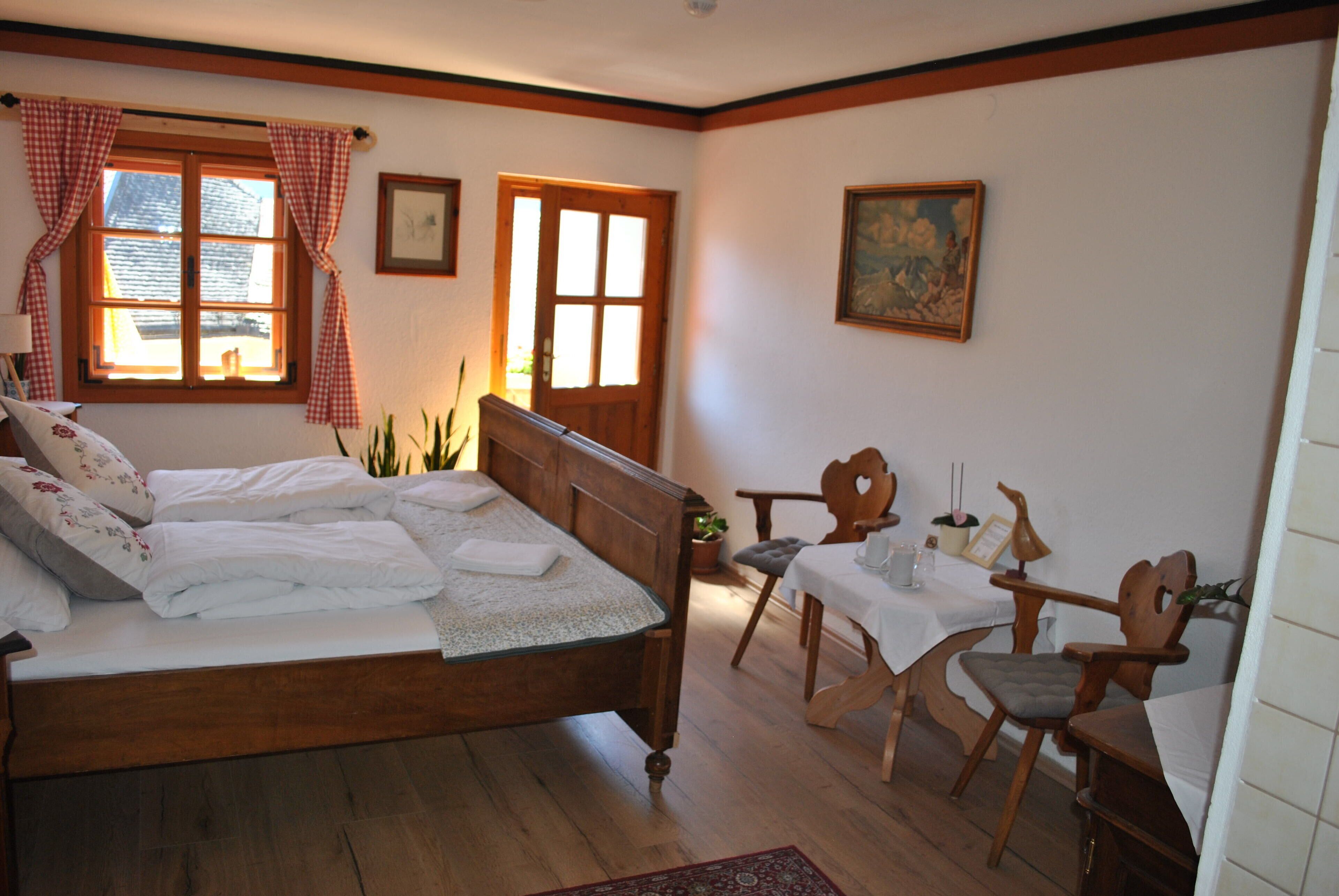 Un dormitorio con cama de madera, una mesa de comedor, dos sillas, una ventana con cortinas y un cuadro en la pared.